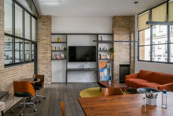 Le Loft : salon avec canapé, grande table en bois, bibliothèque et écran, sous une verrière d'atelier
